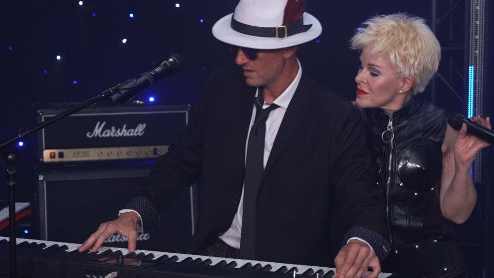 Kelly Corsino leaning in beside the keyboard player in a white fedora