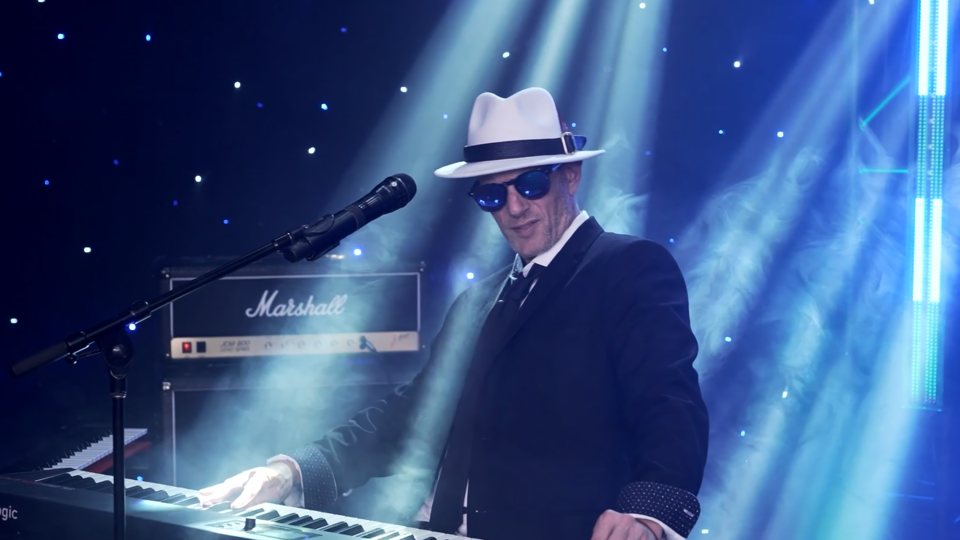 Keyboard player in a white fedora and sunglasses in beams of stage light